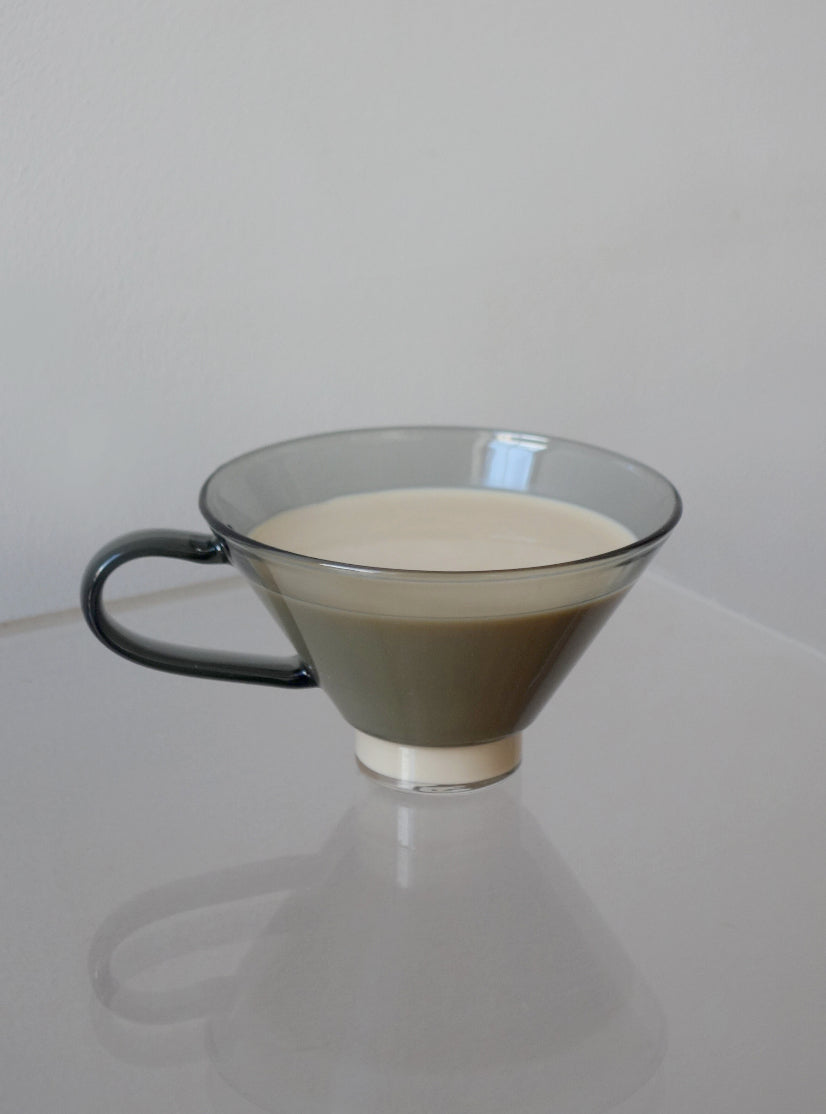 Grey glass cup with a black handle containing coffee with milk on a white surface.